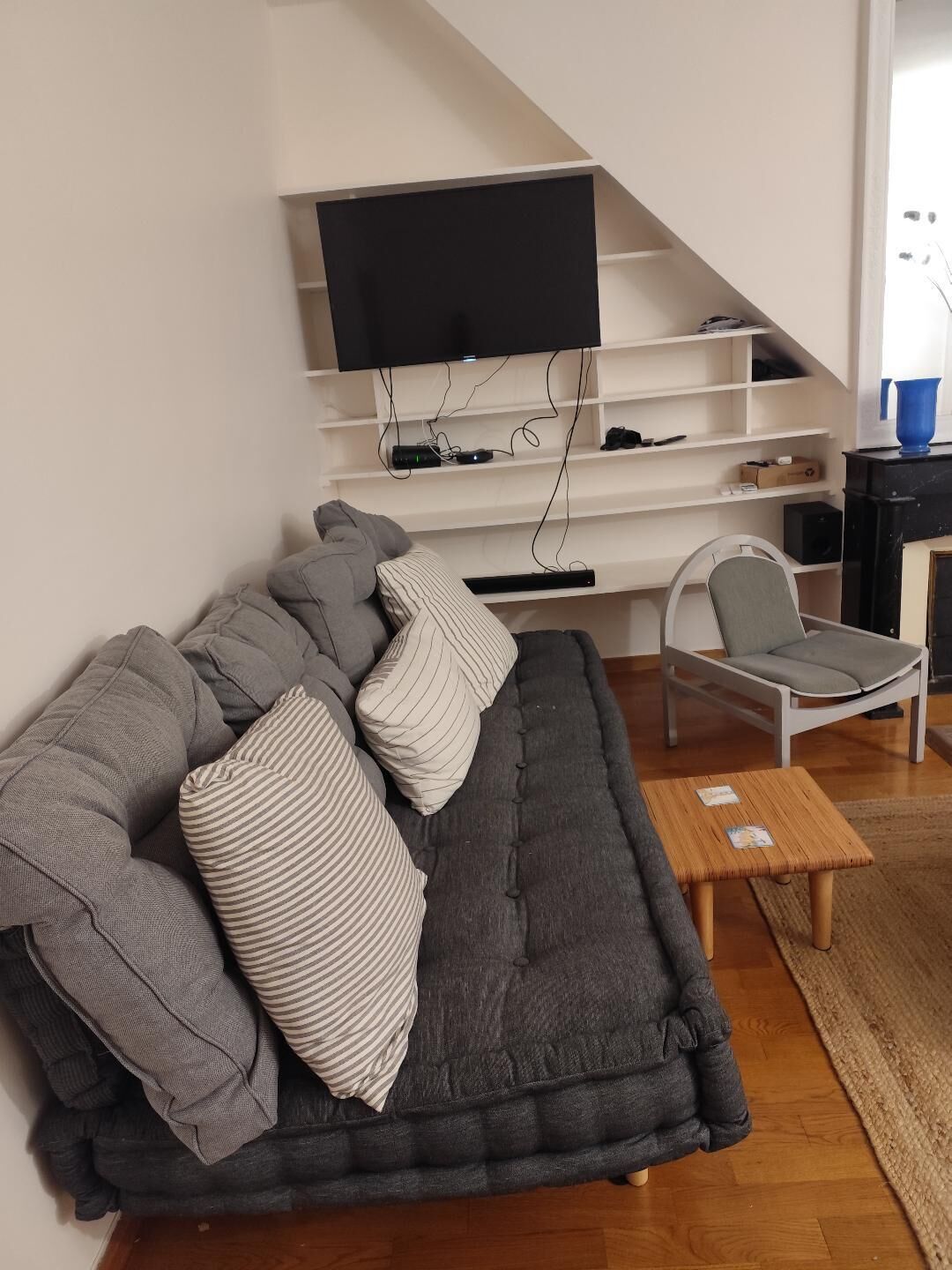 Ein Wohnzimmer mit grauem Sofa, gestreiften Kissen, einem kleinen Holztisch, einem weißen Sessel und einem an der weißen Wand befestigten Fernseher.
