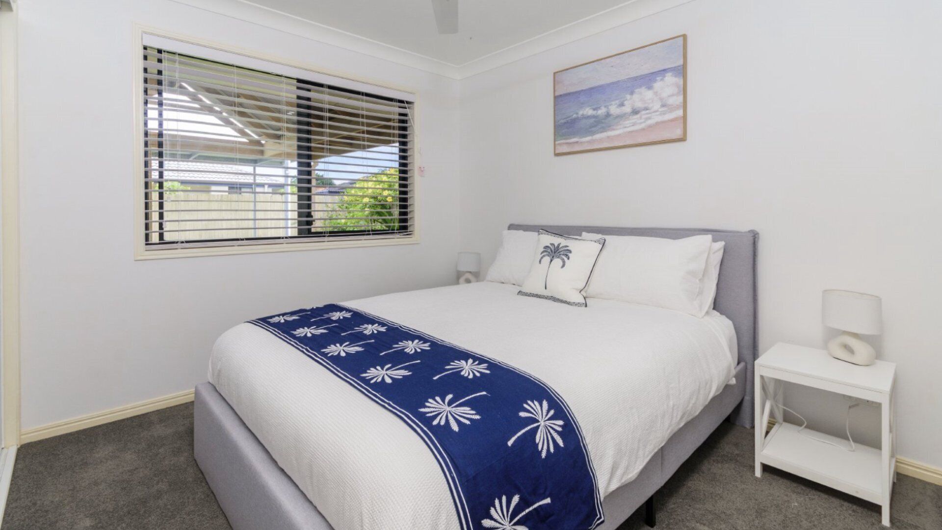 A bedroom with a bed, a nightstand, a lamp, a window with blinds, and a painting on the wall.