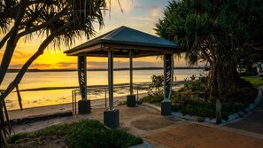A gazebo with a metal roof and concrete base, situated near a beach with a view of the sunset over the water.