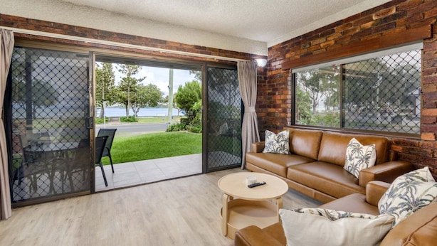 A room with a brown leather sofa, a round coffee table, and a sliding glass door leading to a patio with a lawn and a view of a body of water. - Fairweather Dreaming on Welsby (BONGAREE)