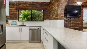 A kitchen with white cabinets, a stainless steel dishwasher, and a brick wall. - Fairweather Dreaming on Welsby (BONGAREE)