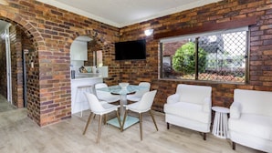 A dining area with a brick wall, a TV mounted above a counter, and white furniture. - Fairweather Dreaming on Welsby (BONGAREE)