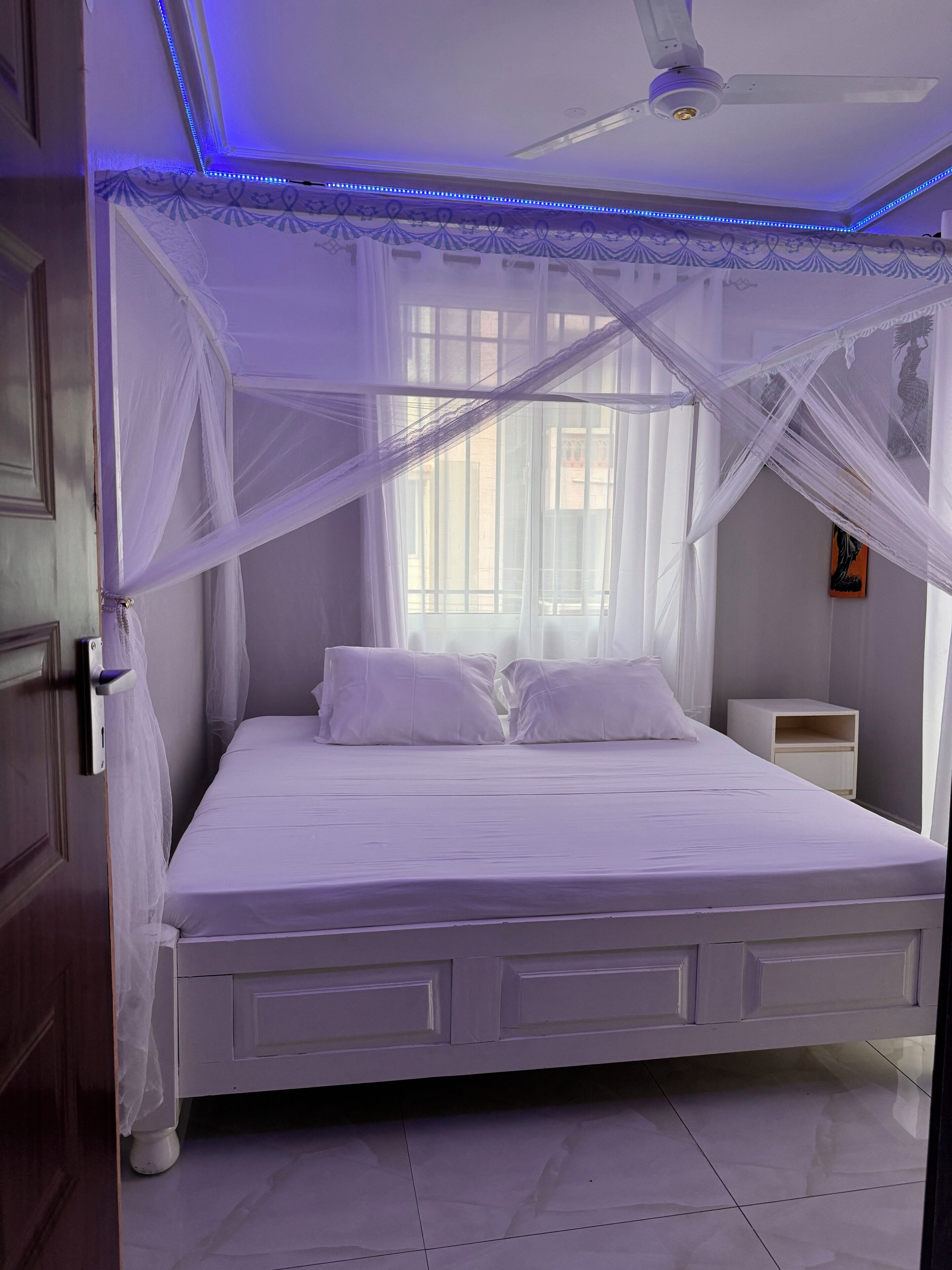 A bedroom with a bed, a nightstand, a ceiling fan, and a window with sheer curtains.