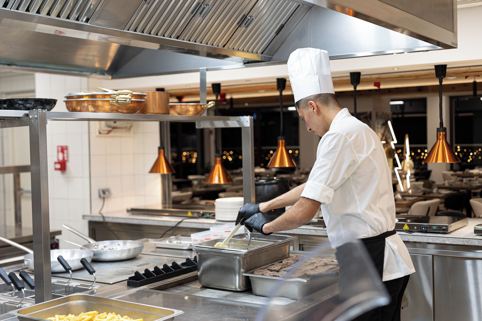 A chef in a professional kitchen with stainless steel equipment, copper pots, and modern pendant lighting.