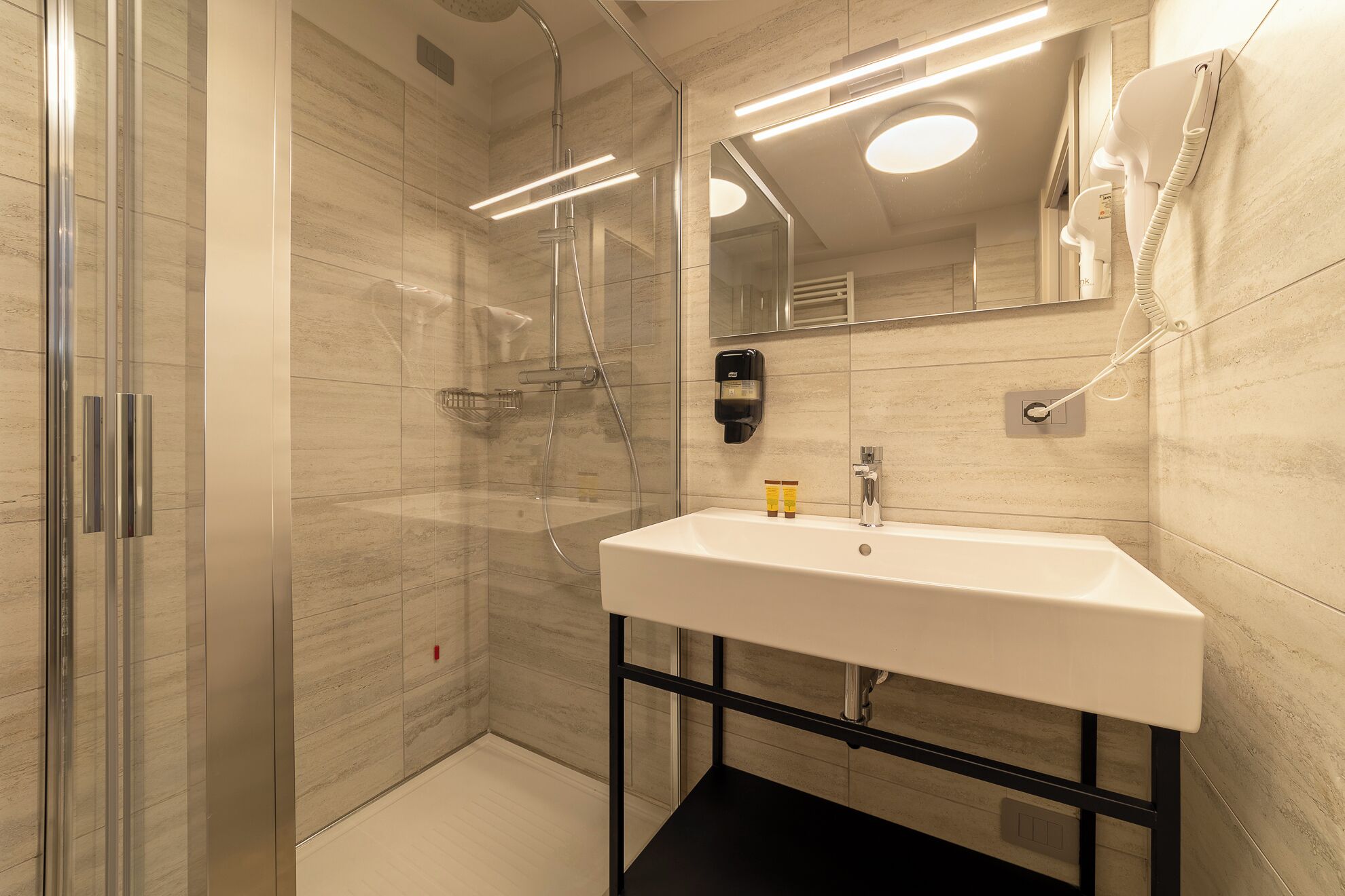 Une salle de bain moderne dotée d’une douche à parois de verre, d’un lavabo blanc à encadrement noir et d’un miroir avec éclairage intégré.