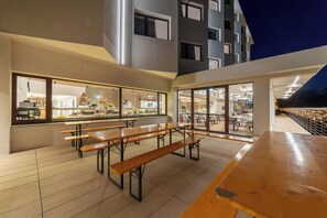 An outdoor dining area with wooden benches and tables, large windows, and a view of the building's interior.