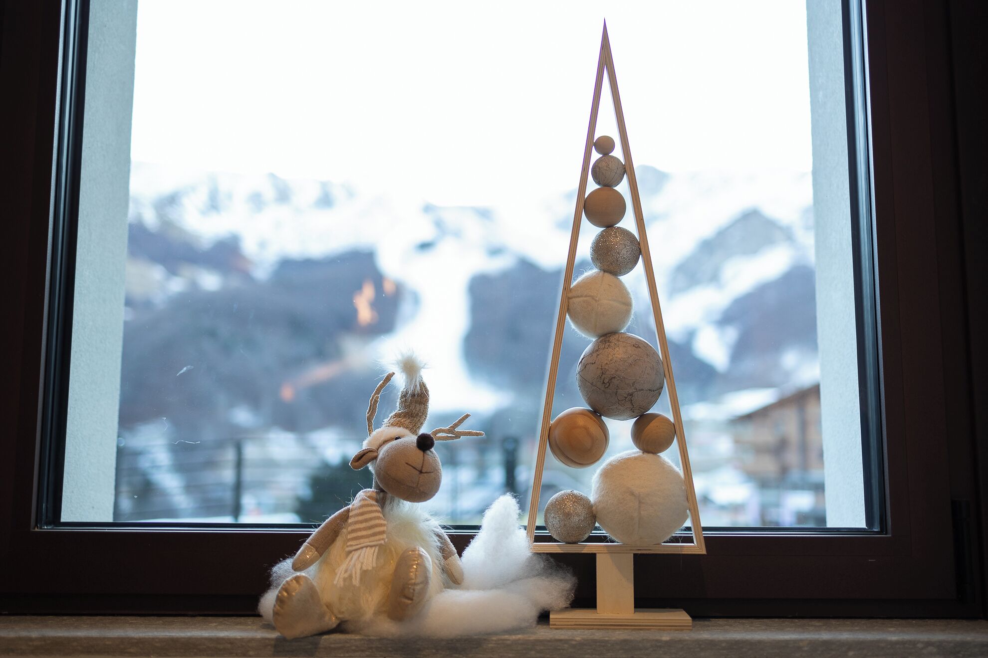 A window with a view of snow-capped mountains, a small wooden Christmas tree, and a plush toy.