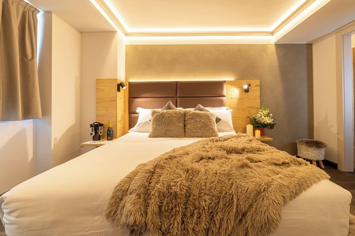 A hotel room with a large bed, a wooden headboard, a fluffy throw blanket, and a nightstand with a bottle and glasses.