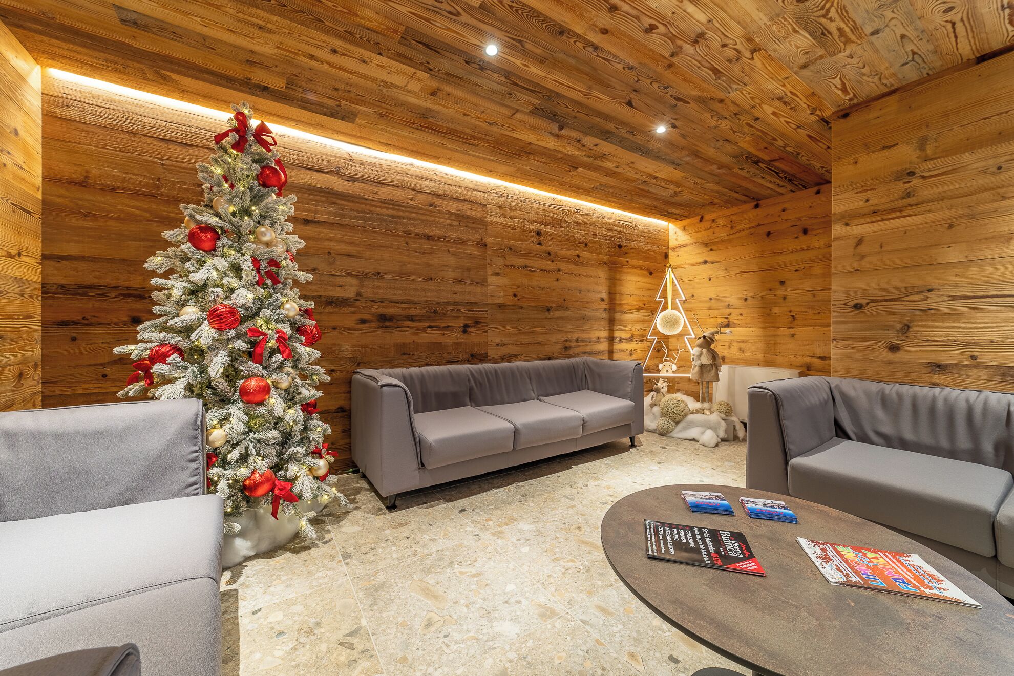 Interior acogedor de una cabaña de madera con un árbol de Navidad decorado, dos sofás grises, una mesa de centro con revistas y un techo de madera.