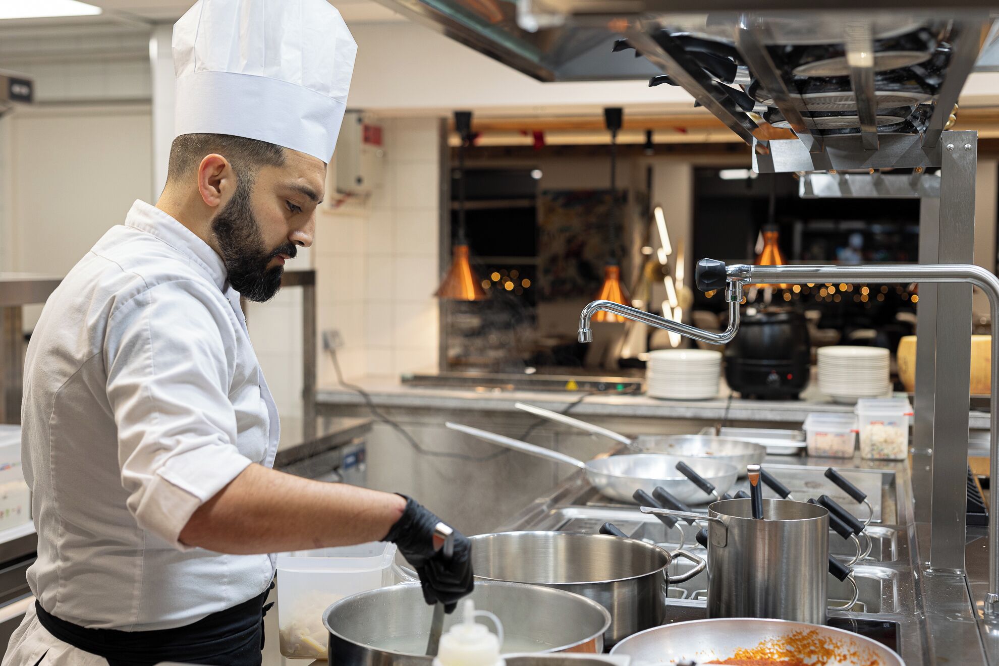 A chef in a professional kitchen with stainless steel cookware, a sink, and overhead cooking equipment.
