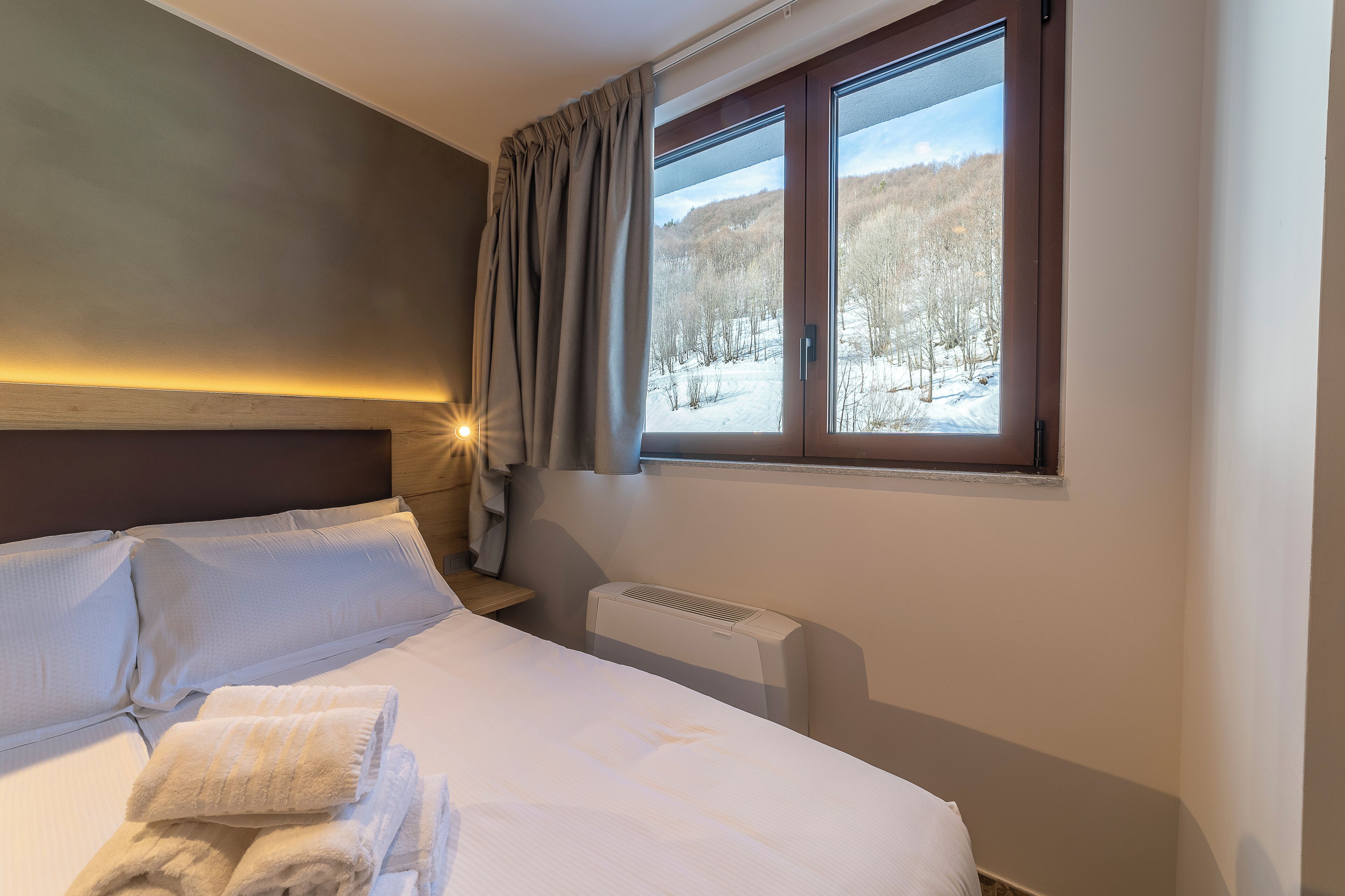 Habitación de hotel con cama, ropa de cama blanca, ventana con vista a un paisaje nevado, calefactor y luz nocturna.