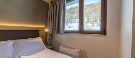 Habitación de hotel con cama, ropa de cama blanca, ventana con vista a un paisaje nevado, calefactor y luz nocturna.