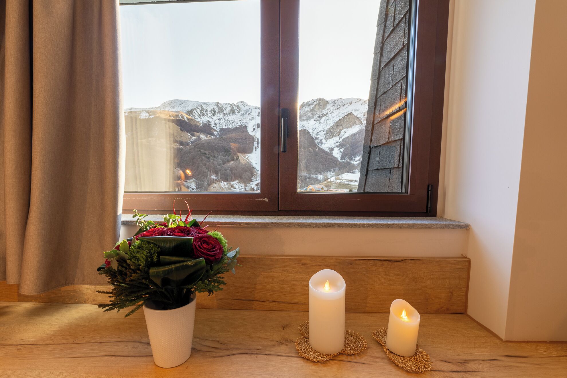 A window with a view of snow-capped mountains, a potted plant with red flowers, and two candles on a wooden ledge.