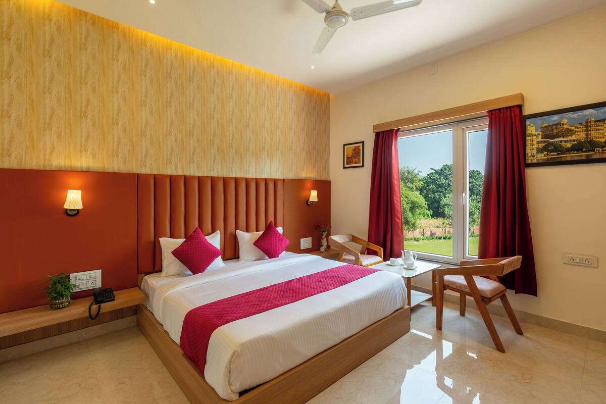 A hotel room with a large bed, a desk, and a chair. There is a window with curtains, a ceiling fan, and a wall-mounted lamp.