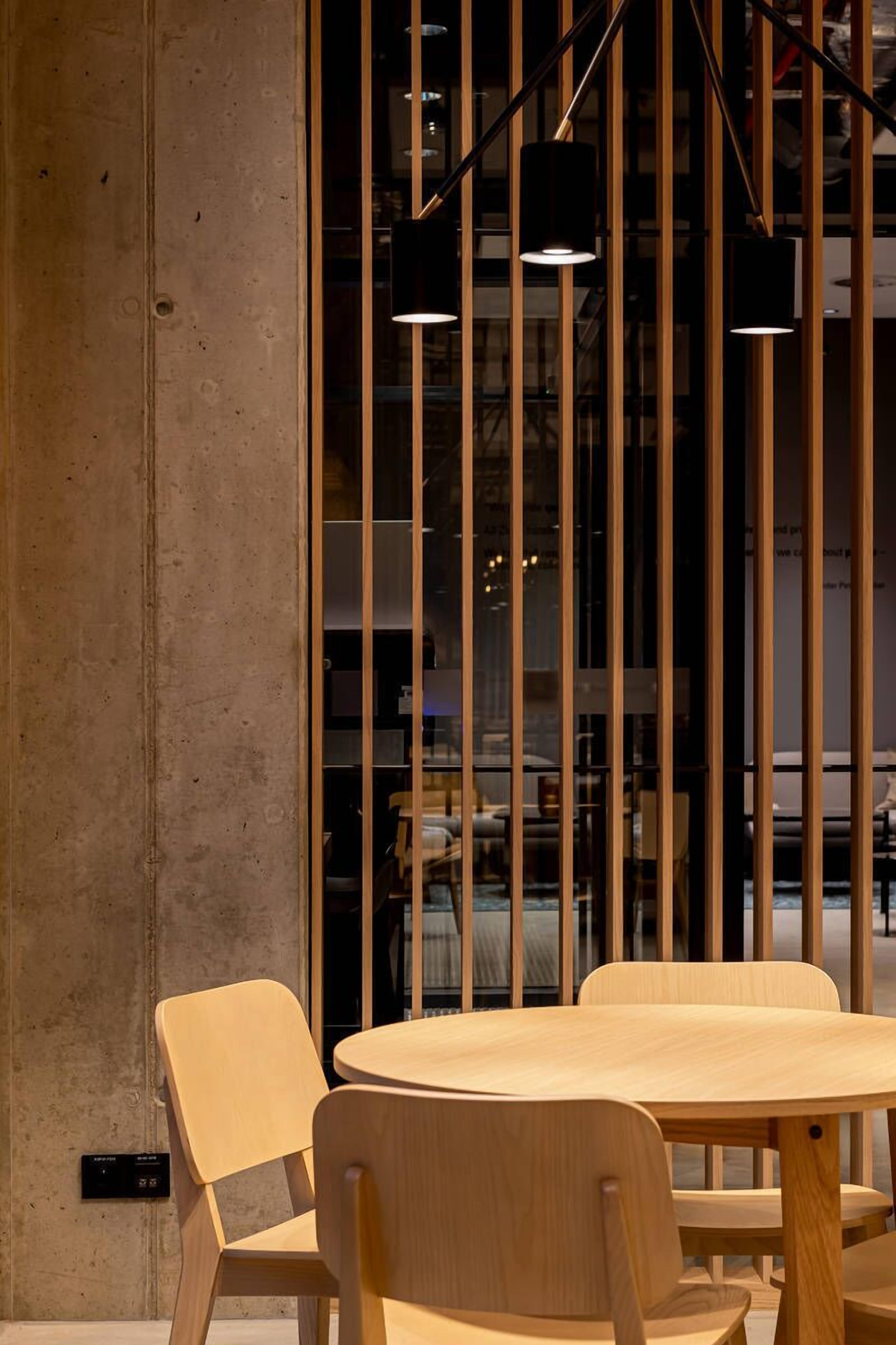 A modern interior with a round wooden table and chairs, a concrete wall, and vertical wooden slats with hanging lights.