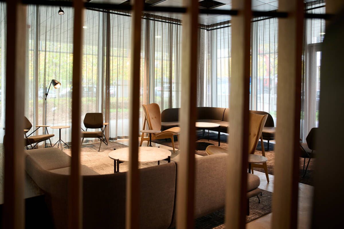 Een moderne loungehoek met diverse zithoeken, waaronder banken en fauteuils, gerangschikt rond een centrale salontafel. Grote ramen met dunne gordijnen laten natuurlijk licht binnen.
