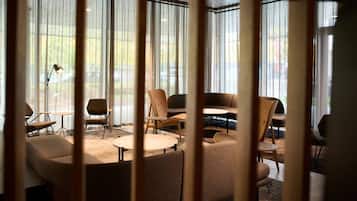 Een moderne loungehoek met diverse zithoeken, waaronder banken en fauteuils, gerangschikt rond een centrale salontafel. Grote ramen met dunne gordijnen laten natuurlijk licht binnen.