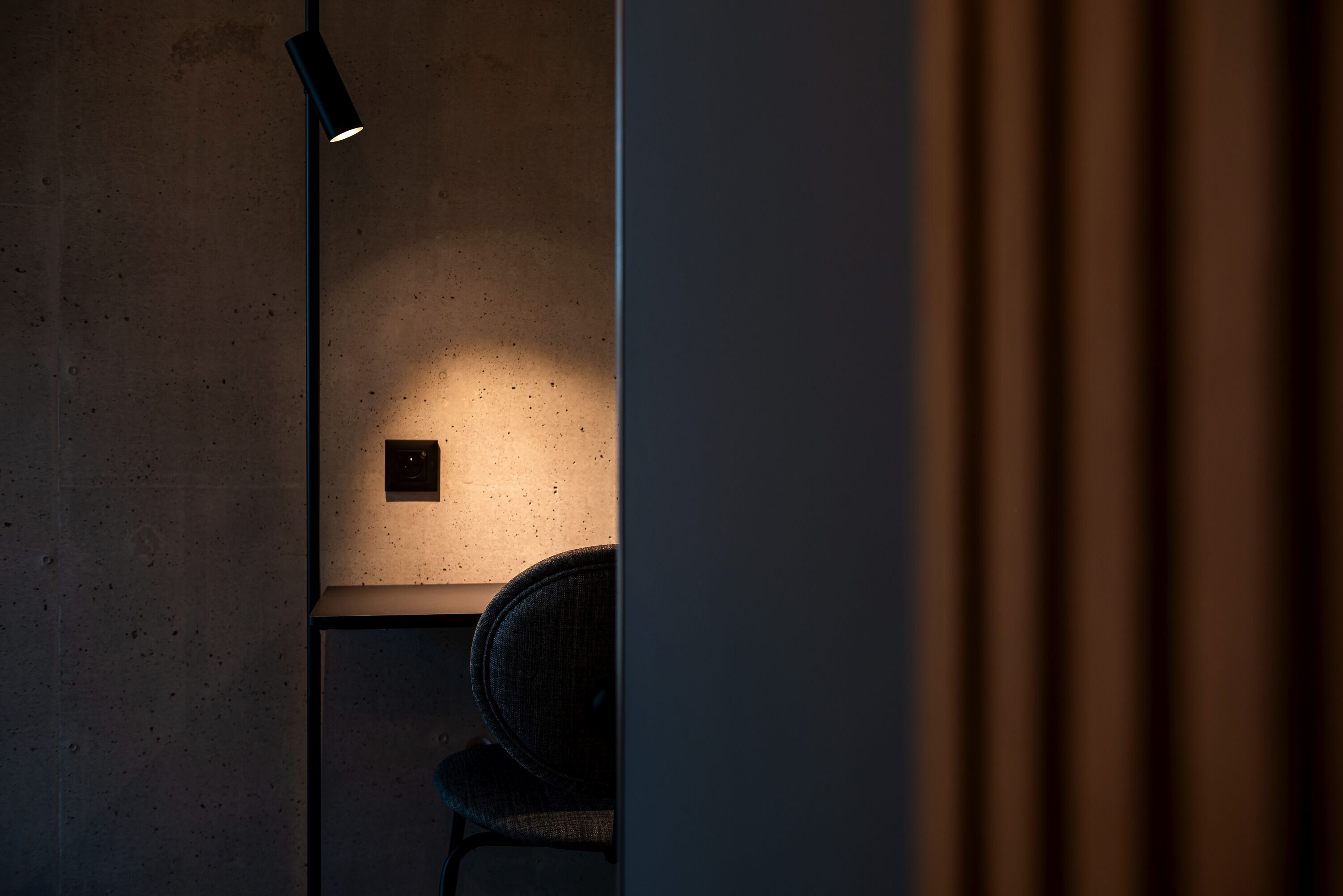 A dimly lit room with a single chair and a small table, a wall-mounted light, and a partial view of a curtain.