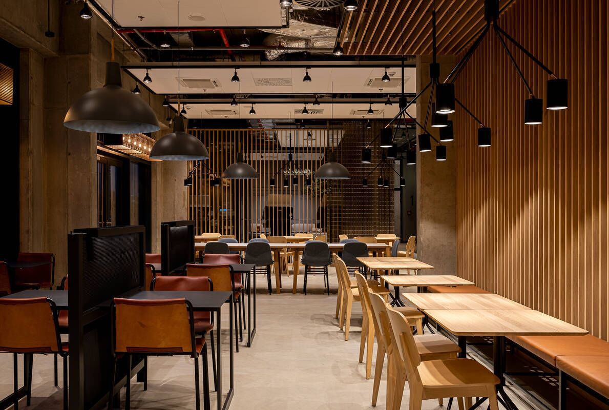 Un interior de restaurante moderno con largas mesas y sillas de madera, iluminación colgante y un techo alto con conductos expuestos.