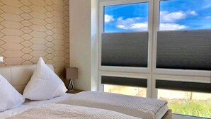 A bedroom with a bed, white bedding, a nightstand, a lamp, and a window with blinds.