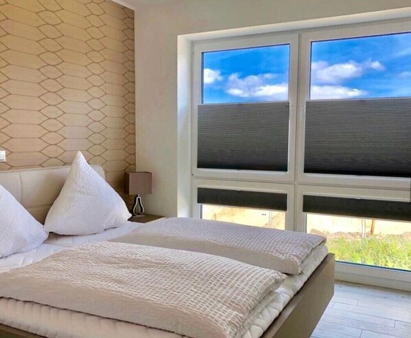 Un dormitorio con una cama, ropa de cama blanca, una mesita de noche, una lámpara y una ventana con persianas.