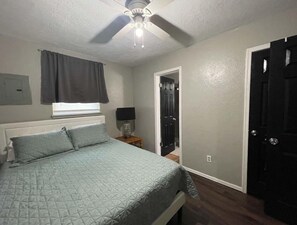Une chambre à coucher comprenant un lit, un ventilateur de plafond, une table de chevet avec une lampe, une porte et une fenêtre avec des rideaux.