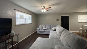 Una sala de estar con un sofá modular, un televisor sobre un mueble bajo, un ventilador de techo y una puerta.