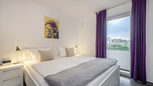 Et moderne soveværelse med en stor seng, et natbord med en lampe, et sengebord og et vindue med lilla gardiner.