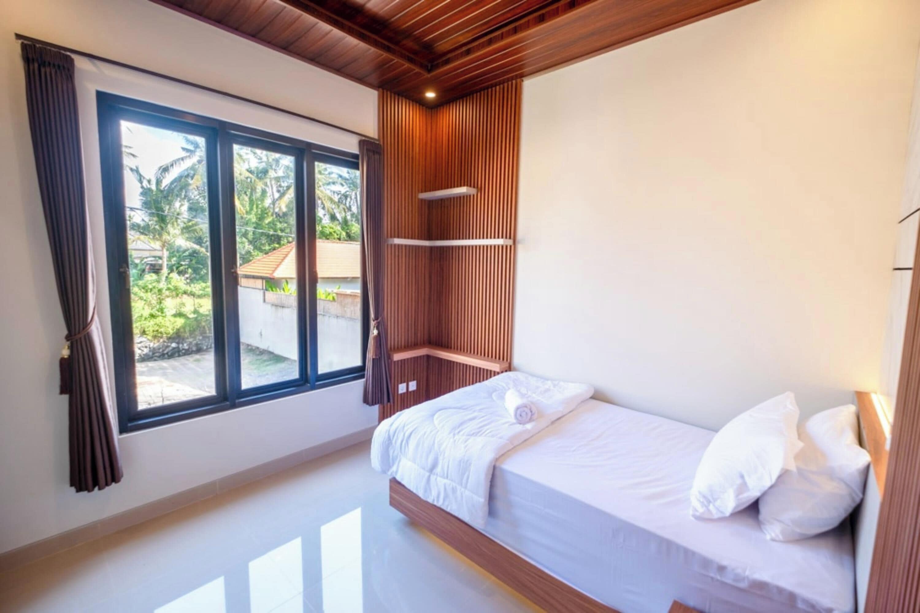 A modern bedroom with a wooden ceiling, a large window, a bed with white bedding, and a view of greenery outside.