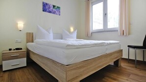 A modern bedroom with a wooden bed frame, white bedding, a nightstand with drawers, a chair, and a window with curtains. - Appartement Hafenflair am Plauer See (Plau am See)