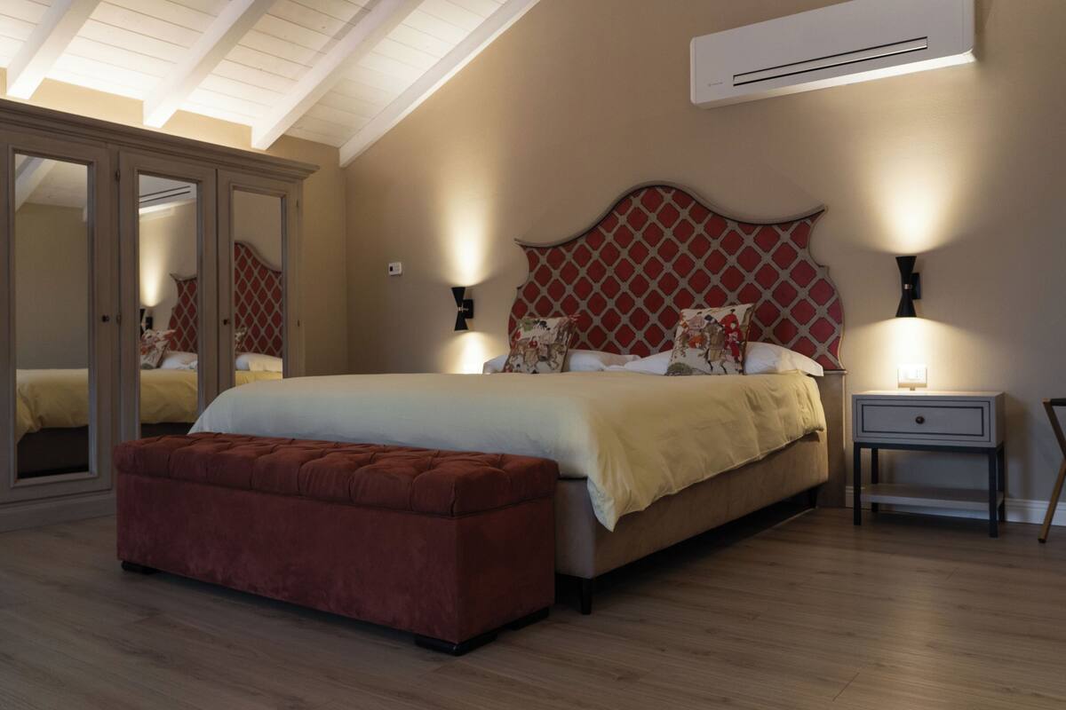 Quarto com cama grande, um banquinho de madeira, mesa de cabeceira e um espelho.