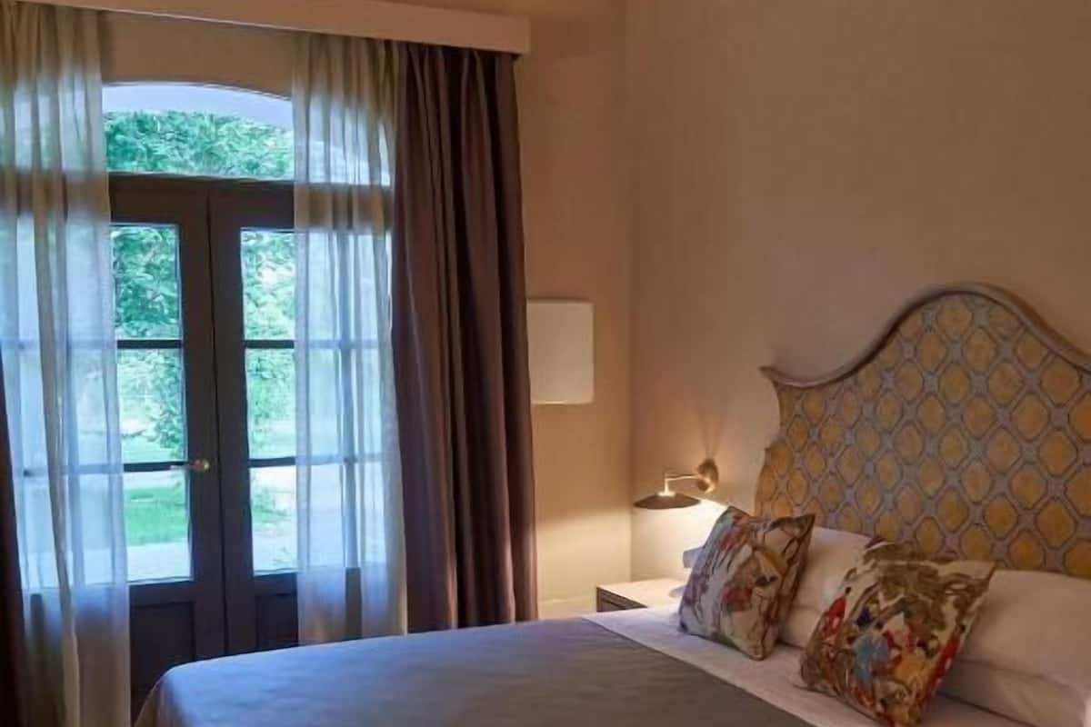 Quarto com cama grande, mesa de cabeceira, luminária e vista para o verde pela janela.