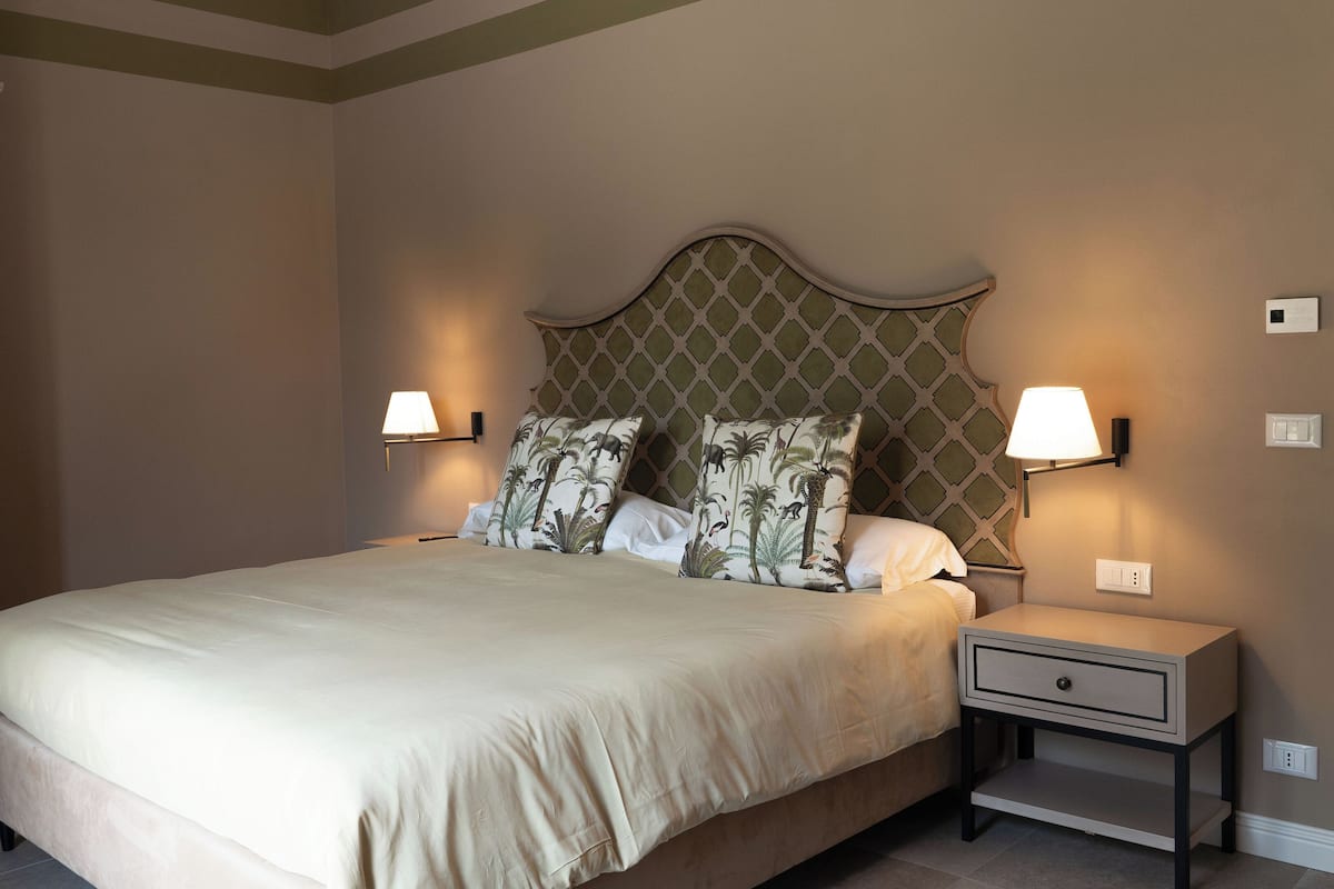 Cama bem arrumada com cabeceira grande, duas luminárias de cabeceira e mesa de cabeceira com gaveta.