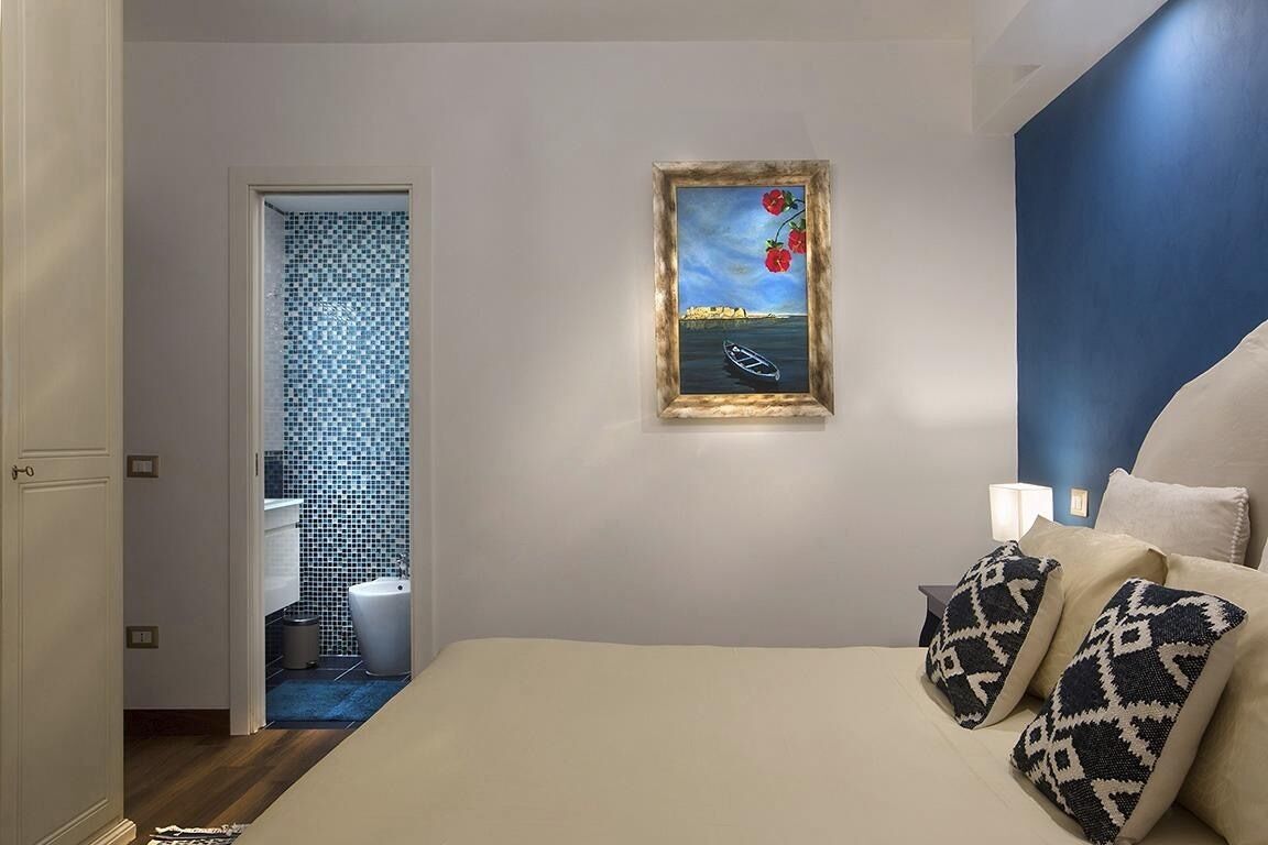 Une chambre à coucher avec un lit, des oreillers, une table de chevet, un tableau encadré et une salle de bain visible à travers une porte ouverte.