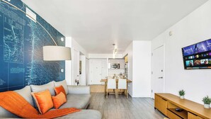 Un salon moderne avec une grande carte accrochée au mur, un canapé orné de coussins orange, un coin repas et un téléviseur diffusant un film.