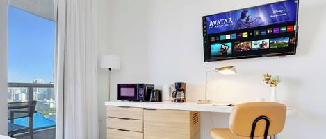 A hotel room with a desk, a flat-screen TV displaying a streaming service, a microwave, a coffee maker, and a view of a cityscape.