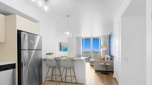 A modern kitchen with a stainless steel refrigerator, two bar stools, a dining area with a table and chairs, a sofa, and a view of the city through large windows. - Modern 2BR 2BA Oasis & pool (Hallandale Beach)
