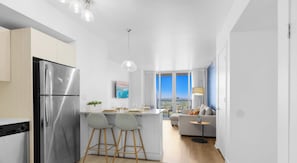 A modern kitchen with a stainless steel refrigerator, two bar stools, a dining area with a table and chairs, a sofa, and a view of the city through large windows. - Modern 2BR 2BA Oasis & pool (Hallandale Beach)