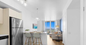 Een moderne keuken met een roestvrijstalen koelkast, twee barkrukken, een eethoek met tafel en stoelen, een bank en een uitzicht op de stad door grote ramen.
