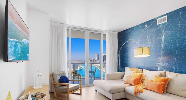 A modern living room with a large TV, a sofa with orange cushions, a wooden chair, and a view of the city through large windows.