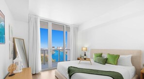 A hotel room with a large bed, a nightstand, a lamp, a view of the city and water through the window.