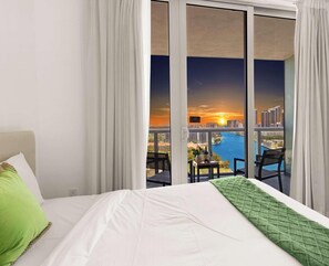 A hotel room with a bed, a green pillow, a view of a cityscape and river at sunset through large windows.
