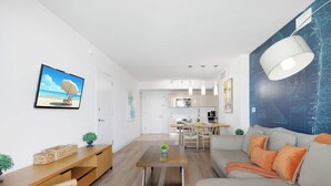A modern living room with a sofa, coffee table, and TV. A kitchen is visible in the background. - Modern Resort - Pool & Amenities (Hallandale Beach)