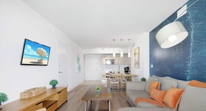 A modern living room with a sofa, coffee table, and TV. A kitchen is visible in the background.