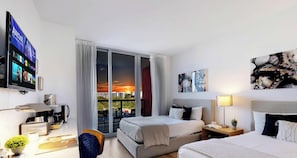 A hotel room with a bed, desk, coffee machine, TV, and a balcony with a sunset view.