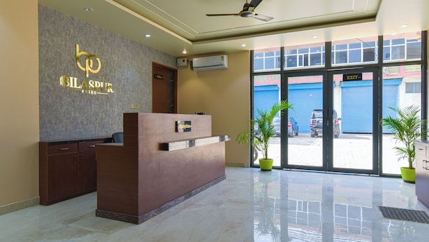 A hotel reception area with a reception desk, a ceiling fan, and a large window with an exit sign. - Hotel Bilaspur Pride (Gurugram)