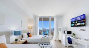 A hotel room with a bed, bedside table, lamp, TV, desk, chair, and a view of the city. - Studio with Amazing Views, pool and gym (Hallandale Beach)