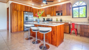 A kitchen with wooden cabinets, a stainless steel refrigerator, a granite countertop, and a large island with bar stools. - Gated Community-Privacy, Ocean Views-pool-Ojochal-sleep 9 (Ojochal)