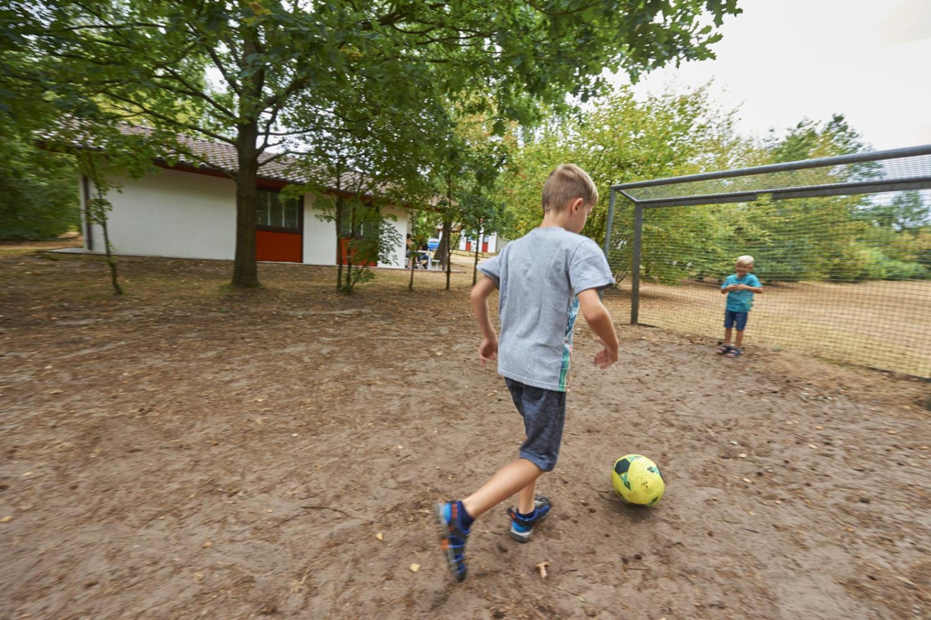 Un garçon joue au football dans un espace extérieur, avec un filet, des arbres et un bâtiment en arrière-plan.