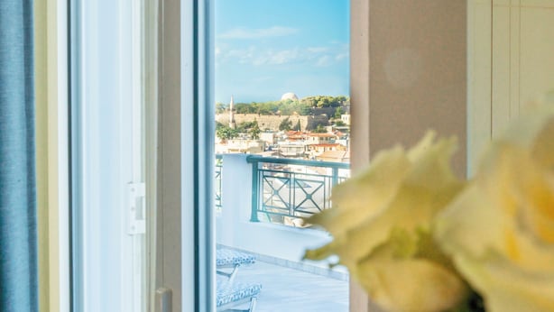 Vista a través de una ventana con una flor blanca en primer plano, un balcón con barandilla y una ciudad distante con edificios y una estructura en forma de cúpula.